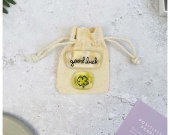 May include: A beige drawstring bag with two glass pebbles. The top pebble has the words "good luck" in black script. The bottom pebble is square with a green four-leaf clover design. A card with the words "POSITIVITY PEBBLE" is visible.