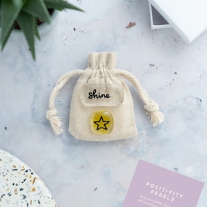 May include: A small white drawstring bag containing a yellow glass pebble. The pebble features a black star and the inscription "Shine". A card with the text "Positivity Pebble" is also included. Keep your pebble close to hand and remember its message as you hold it right.