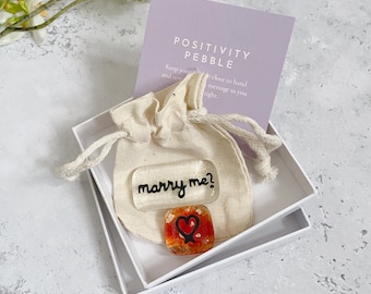 May include: A white gift box contains a small beige drawstring bag and two glass pebbles. One pebble says "marry me?" in black script. The other is square with a red heart design. A purple card reads "POSITIVITY PEBBLE."