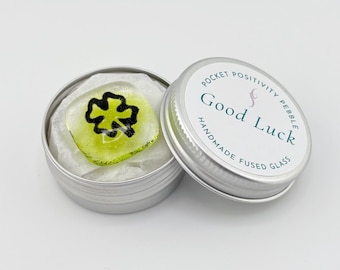 May include: A handmade fused glass pocket positivity pebble with a four-leaf clover design in a small silver tin. The pebble is lime green with a black clover. The tin lid reads "Good Luck".