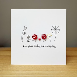 May include: A white greeting card featuring two red circular embellishments representing birds, a small red heart, and black line drawings of flowers and a dandelion. The text "On your Ruby anniversary" is printed in black script at the bottom centre.
