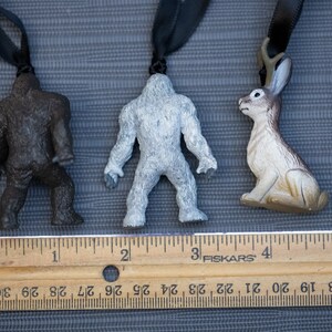 May include: Three small plastic figurines hanging from black cords. The figurines are a brown Bigfoot, a white Bigfoot, and a brown rabbit with antlers. A wooden ruler is below the figurines with the numbers 1 through 6 visible.