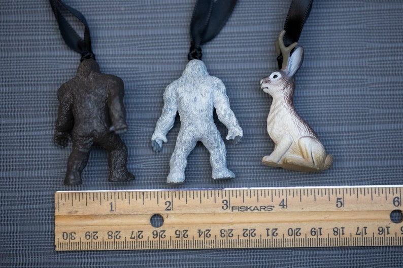 May include: Three small plastic figurines hanging from black cords. The figurines are a brown Bigfoot, a white Bigfoot, and a brown rabbit with antlers. A wooden ruler is below the figurines with the numbers 1 through 6 visible.