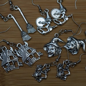 May include: A collection of silver-toned Halloween-themed earrings. Designs include witch hats, witch on a broom, witch on a drum, and a haunted house. The earrings are displayed on a wooden surface.
