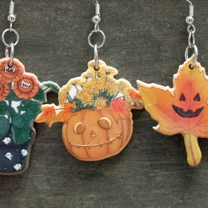 Op de afbeelding: Drie Halloween-oorbellen met zilveren haakjes. De eerste oorbel heeft een plant in een pot met drie oranje bloemen met tanden. De tweede oorbel heeft een jack-o'-lantern met een gele bloemschikking. De derde oorbel heeft een esdoornblad met een jack-o'-lantern gezicht.