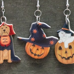 May include: Three Halloween-themed earrings featuring a pumpkin with a witch's hat, a pumpkin with a cat inside, and a dog wearing a cape and a "Trick or Treat" sign.