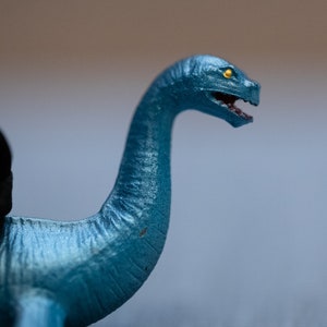 May include: A blue plastic toy dinosaur with a long neck and a yellow eye. The dinosaur is posed with its head tilted to the side and its mouth open.