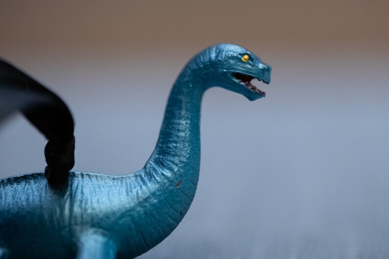 May include: A blue plastic toy dinosaur with a long neck and a yellow eye. The dinosaur is posed with its head tilted to the side and its mouth open.