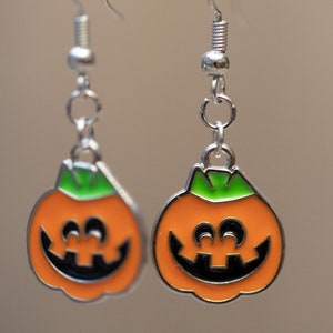 May include: A pair of silver dangle earrings with orange and green pumpkin charms. The pumpkins have black smiling faces.