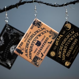 May include: Three square Ouija board earrings with silver hooks hang from a black branch. One is black with gold lettering, another is tan with black lettering, and the third is black with white lettering. The earrings feature the alphabet, numbers, and the words "Yes", "No", "Ouija", "Happy Halloween", "Trick", "Treat", and "Farewell".