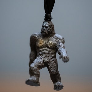 May include: A brown and gray figurine of Bigfoot hanging from a black string. The figurine is detailed with fur and has a serious expression.