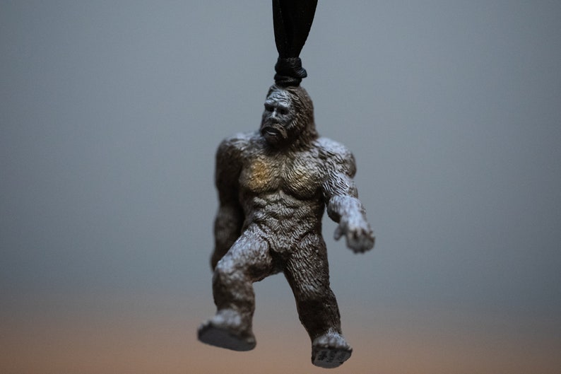 May include: A brown and gray figurine of Bigfoot hanging from a black string. The figurine is detailed with fur and has a serious expression.
