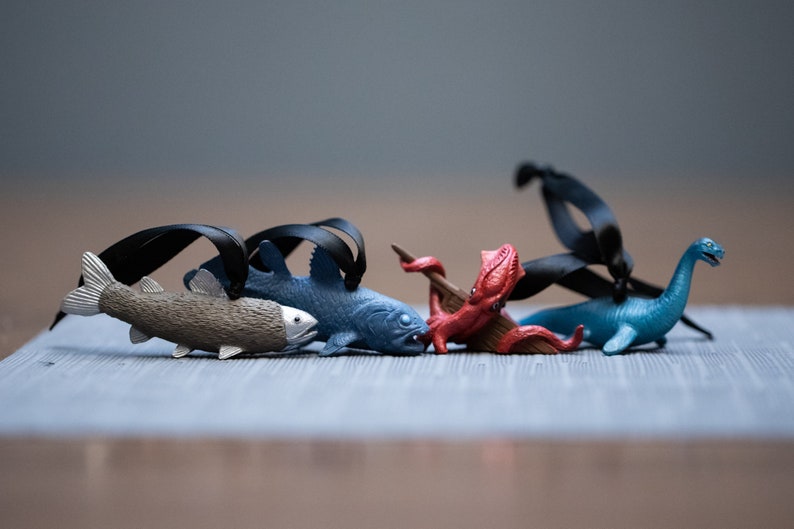 May include: Five colorful plastic sea creature ornaments with black ribbon hangers. The ornaments include a blue fish, a gray fish, a red octopus, and a blue sea serpent.