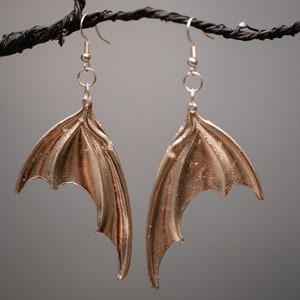 May include: A pair of silver-colored earrings shaped like bat wings. The earrings are hanging from a black branch.