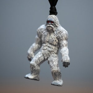 May include: A white, furry figurine of a yeti with red eyes. The figurine is hanging from a black cord.
