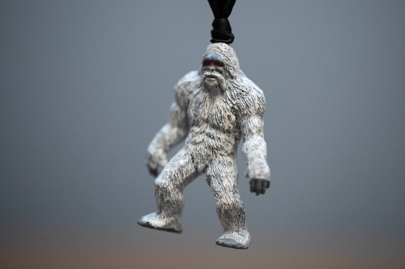 May include: A white, furry figurine of a yeti with red eyes. The figurine is hanging from a black cord.