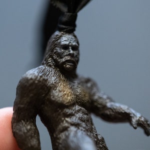 May include: A small, detailed figurine of Bigfoot, a mythical creature, made of dark gray metal. The figurine is holding its arms out, and its face has a surprised expression.