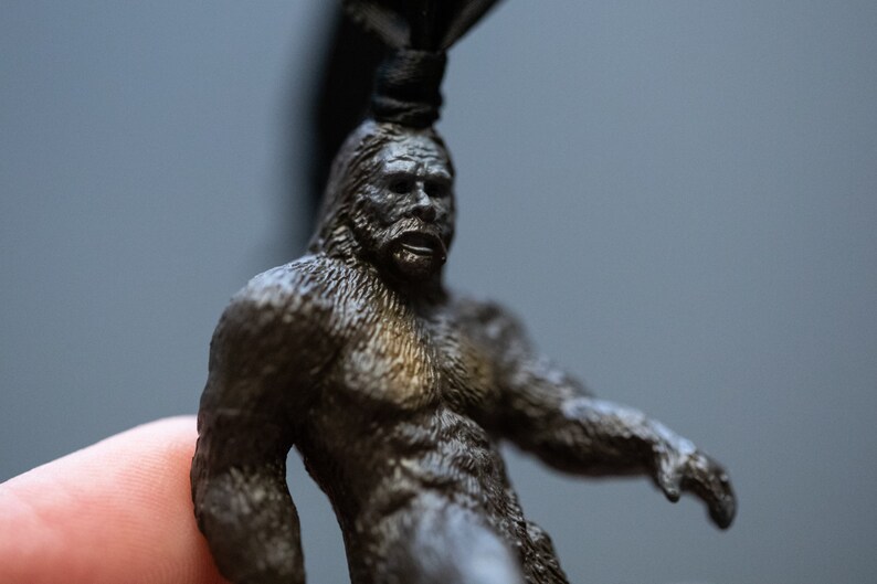 May include: A small, detailed figurine of Bigfoot, a mythical creature, made of dark gray metal. The figurine is holding its arms out, and its face has a surprised expression.