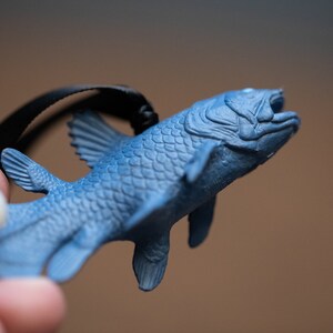 May include: A blue plastic figurine of a coelacanth, a prehistoric fish, hanging from a black cord. The figurine is detailed with scales and fins.