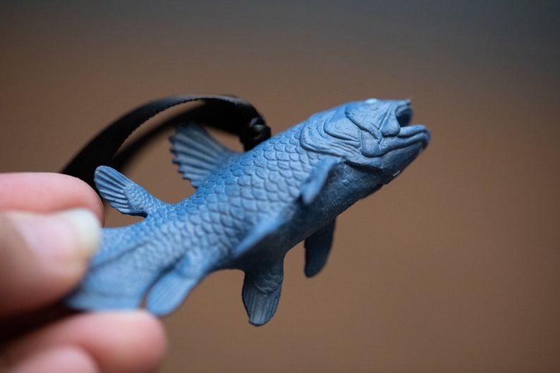 May include: A blue plastic figurine of a coelacanth, a prehistoric fish, hanging from a black cord. The figurine is detailed with scales and fins.