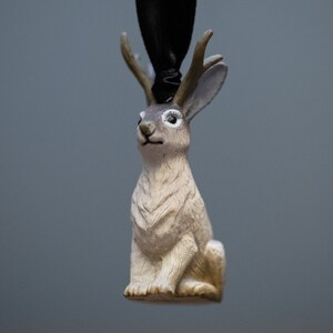 May include: A small, white rabbit figurine with brown markings and antlers. The figurine is made of plastic and has a black ribbon attached to the top of its head.