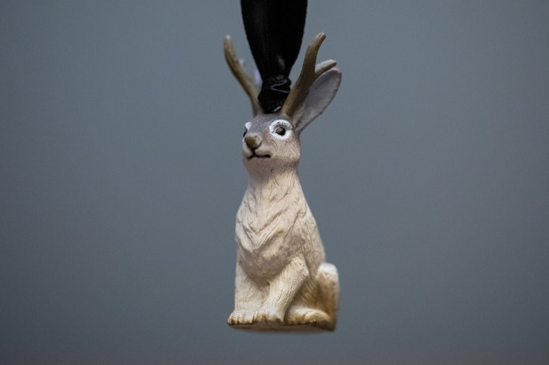 May include: A small, white rabbit figurine with brown markings and antlers. The figurine is made of plastic and has a black ribbon attached to the top of its head.