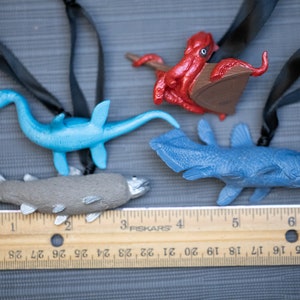 May include: Four plastic sea creature ornaments hanging from black cords. The ornaments are a blue plesiosaur, a gray fish, a blue coelacanth, and a red octopus. A wooden ruler with black markings is in the bottom of the image.