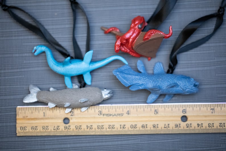 May include: Four plastic sea creature ornaments hanging from black cords. The ornaments are a blue plesiosaur, a gray fish, a blue coelacanth, and a red octopus. A wooden ruler with black markings is in the bottom of the image.