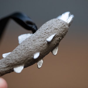 May include: A silver-colored fish charm with a textured surface and a black cord attached. The fish has a slightly open mouth and fins.