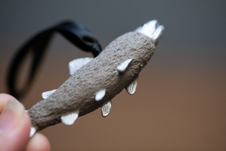 May include: A silver-colored fish charm with a textured surface and a black cord attached. The fish has a slightly open mouth and fins.