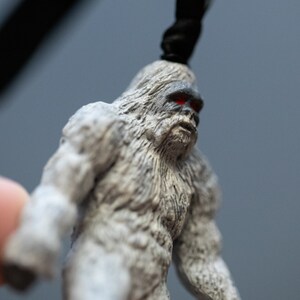 May include: A small, gray, furry figurine of a Bigfoot with red eyes. The figurine is made of plastic and has a detailed texture.