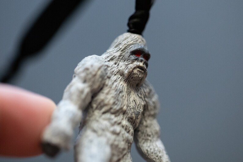 May include: A small, gray, furry figurine of a Bigfoot with red eyes. The figurine is made of plastic and has a detailed texture.
