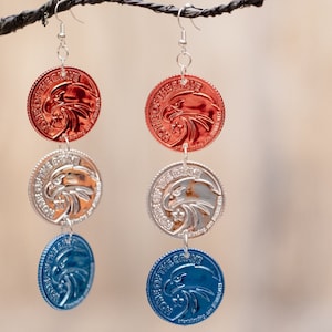 May include: A pair of dangle earrings with three circular charms. The top charm is red with a gold eagle design, the middle charm is silver with a gold eagle design, and the bottom charm is blue with a gold eagle design.