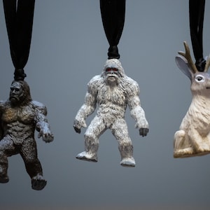 May include: Three small figurines hanging from black cords. The figurines are a brown Bigfoot, a white Bigfoot, and a white rabbit with antlers.