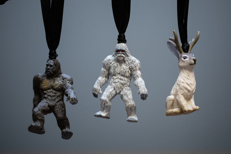 May include: Three small figurines hanging from black cords. The figurines are a brown Bigfoot, a white Bigfoot, and a white rabbit with antlers.