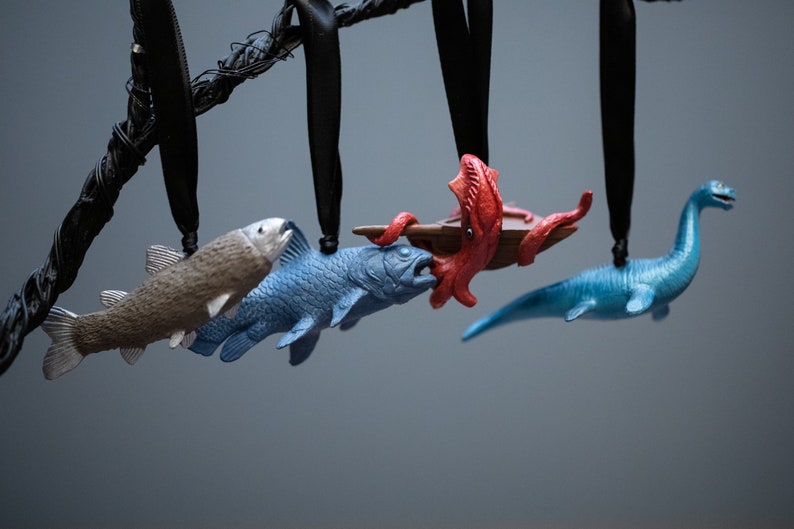 May include: Five hanging ornaments, each depicting a different sea creature. A brown fish, a blue fish, a red octopus, a blue fish, and a blue plesiosaur.