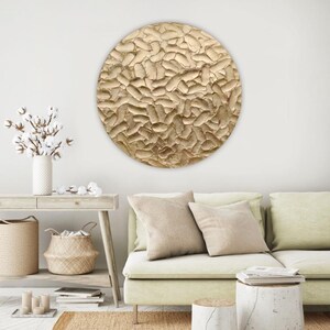 A large, round, textured wall art piece with a gold finish. The artwork has a raised, organic pattern and is mounted on a white wall. The room also features a light green sofa, neutral-toned pillows, and a woven rug.