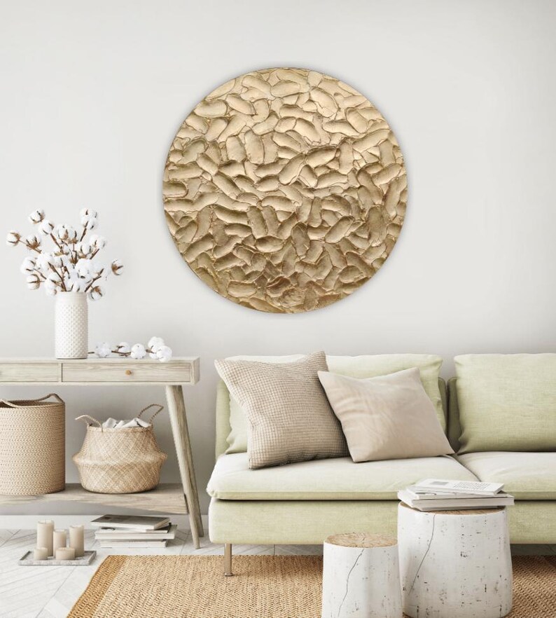 A large, round, textured wall art piece with a gold finish. The artwork has a raised, organic pattern and is mounted on a white wall. The room also features a light green sofa, neutral-toned pillows, and a woven rug.