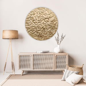 A large, round, textured wall art piece with a gold finish hangs above a light wood cabinet. The art has a raised, organic pattern. A woven lamp, vase, and basket complete the room's decor.