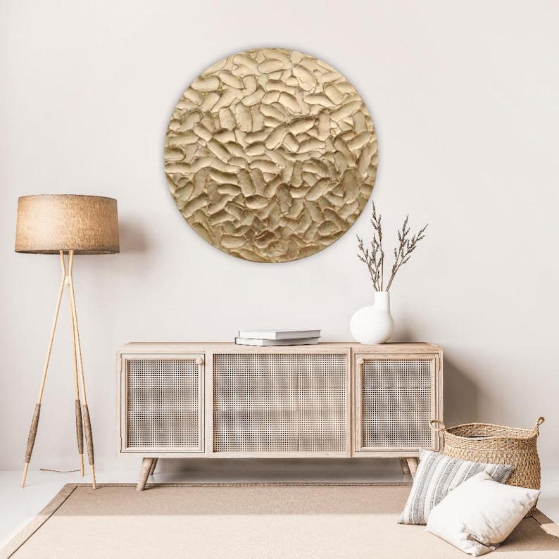A large, round, textured wall art piece with a gold finish hangs above a light wood cabinet. The art has a raised, organic pattern. A woven lamp, vase, and basket complete the room's decor.