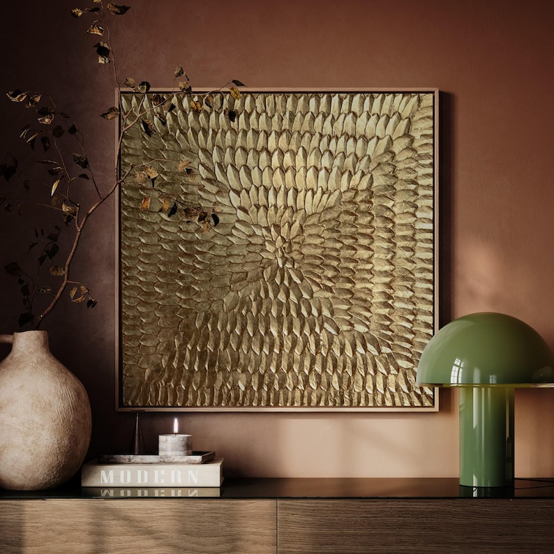 Peut inclure: Une &oelig;uvre d'art murale carr&eacute;e dor&eacute;e avec un motif textur&eacute; en forme de fleur. L'&oelig;uvre est encadr&eacute;e dans un bois clair. Une lampe verte en forme de champignon est pos&eacute;e sur une surface sombre, avec un vase, une bougie et un livre.