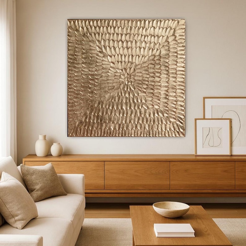 Peut inclure: Une grande &oelig;uvre d'art murale carr&eacute;e de couleur or avec un motif textur&eacute; en relief. L'&oelig;uvre est mont&eacute;e au-dessus d'un meuble en bois dans un salon. Le design pr&eacute;sente un motif central circulaire.