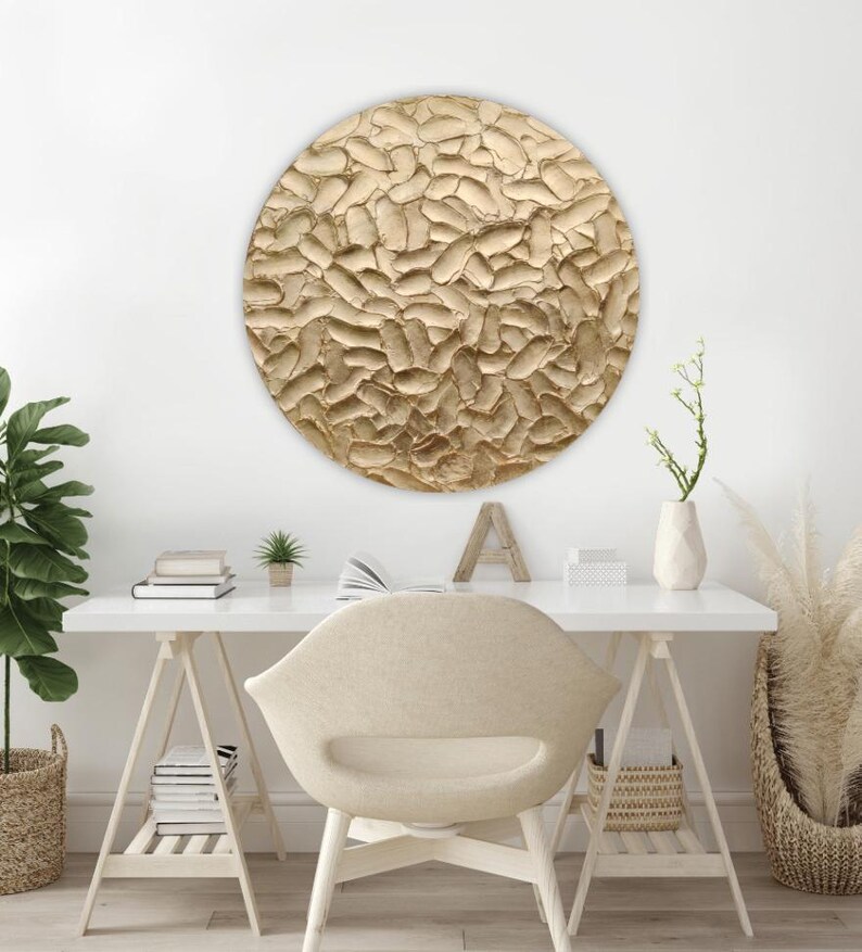 A round, textured wall art piece in shades of gold hangs above a white desk. The artwork has a raised, organic pattern. A white desk, chair, and decorative items complete the modern, neutral-toned office space.
