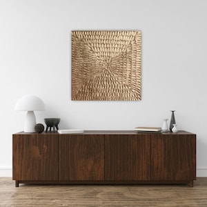 Peut inclure: Une &oelig;uvre d'art murale carr&eacute;e et textur&eacute;e de couleur or est suspendue au-dessus d'un meuble en bois fonc&eacute;. Le meuble comporte quatre portes et est orn&eacute; d'objets d&eacute;coratifs, dont une lampe blanche, un bol noir et de petits vases.