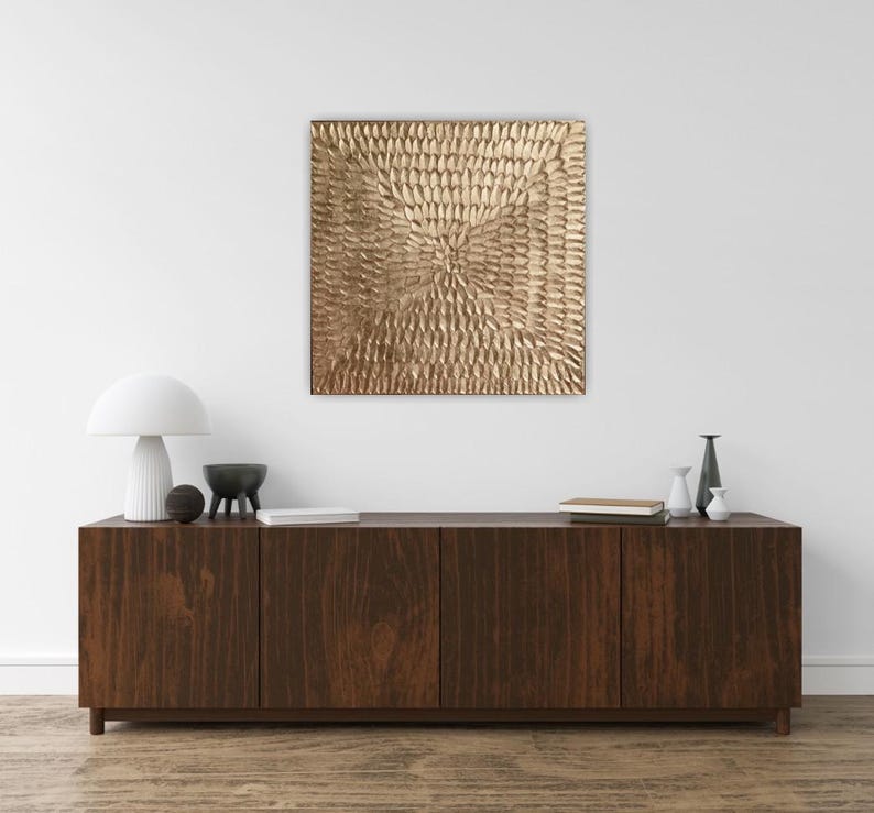 Peut inclure: Une &oelig;uvre d'art murale carr&eacute;e et textur&eacute;e de couleur or est suspendue au-dessus d'un meuble en bois fonc&eacute;. Le meuble comporte quatre portes et est orn&eacute; d'objets d&eacute;coratifs, dont une lampe blanche, un bol noir et de petits vases.