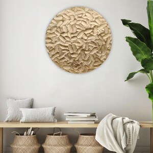 A round, textured wall art piece with a gold finish hangs on a white wall. The artwork has a raised, organic pattern. Below, a wooden bench holds pillows, books, and woven baskets, creating a cozy, neutral-toned interior.