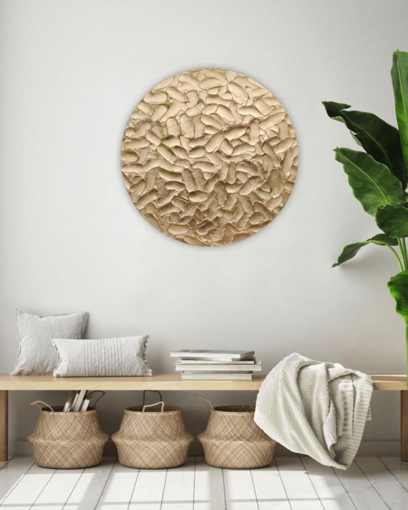 A round, textured wall art piece with a gold finish hangs on a white wall. The artwork has a raised, organic pattern. Below, a wooden bench holds pillows, books, and woven baskets, creating a cozy, neutral-toned interior.