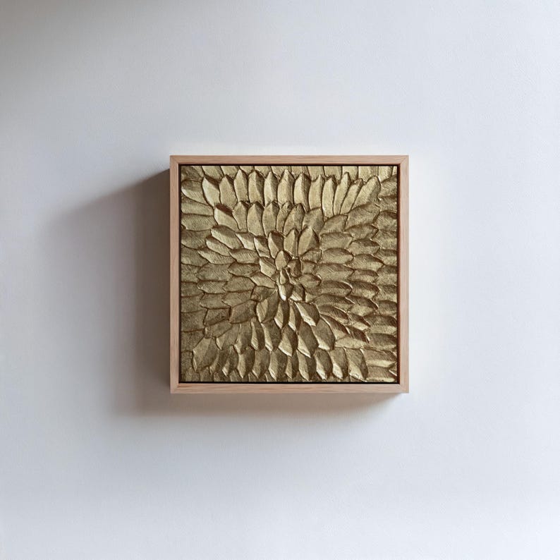 A square abstract painting featuring a heavy 3D hand-sculpted radial petal pattern in metallic gold. The highly textured artwork is displayed in a natural light wood floating frame, hanging against a crisp white wall. Modern botanical sunburst relief decor.
