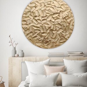 A large, round, textured wall art piece with a gold finish. The artwork has a raised, organic pattern. The piece is mounted above a bed with white pillows and a light pink throw blanket.