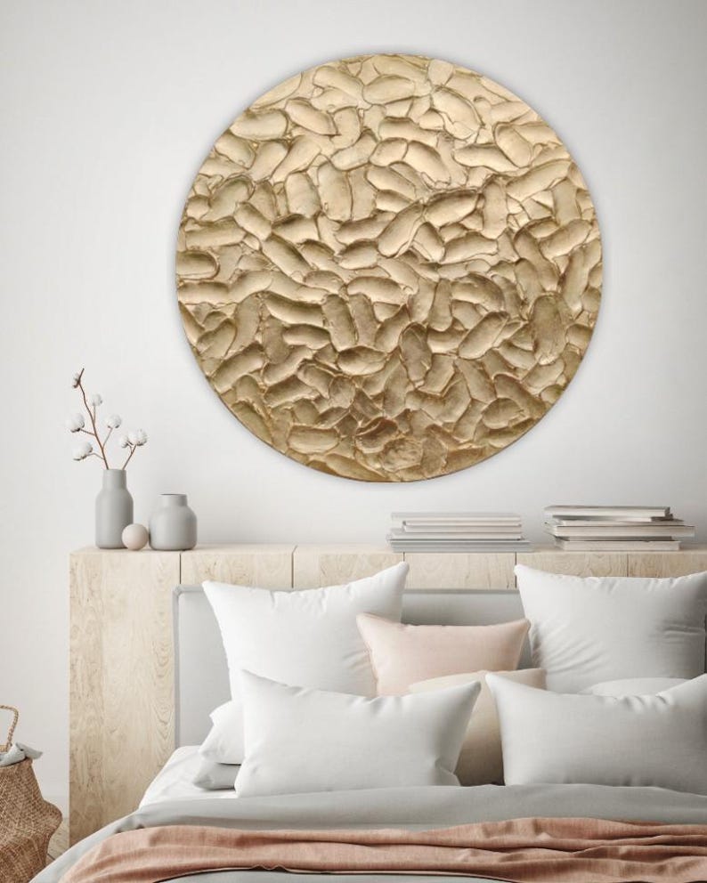 A large, round, textured wall art piece with a gold finish. The artwork has a raised, organic pattern. The piece is mounted above a bed with white pillows and a light pink throw blanket.